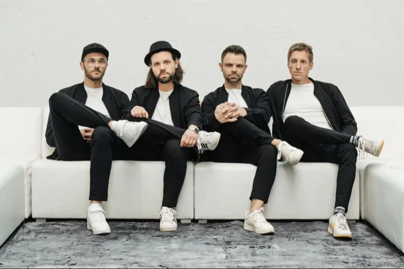 Die vier Musiker der Eventband GOODBEATS in schwarzen Sakkos und weißen Shirts nebeneinander auf einem weißen Sofa