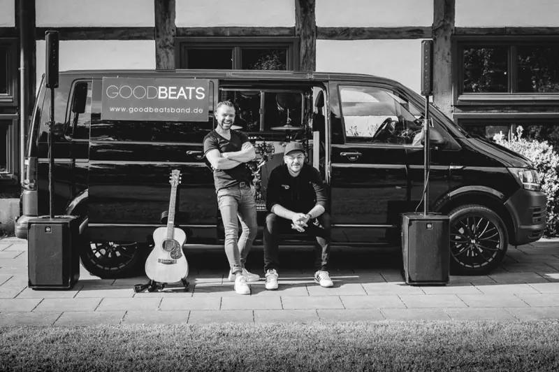 Das Konzert-Taxi: schwarzer Bulli mit GOODBEATS-Banner, davor zwei Musiker, eine Akustikgitarre und zwei Lautsprecher