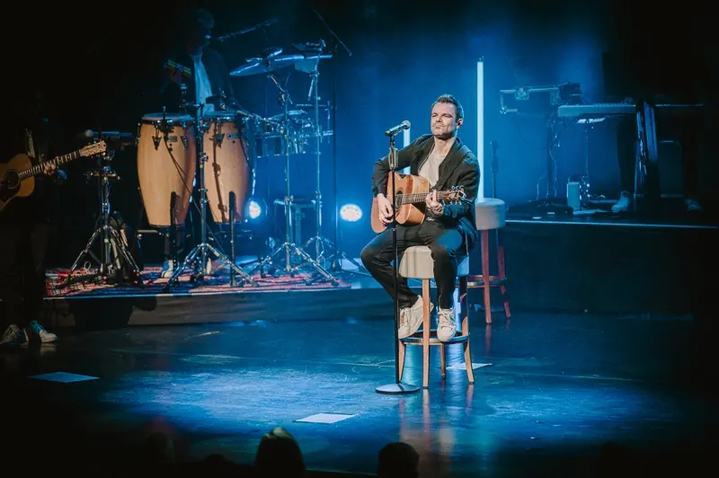 Winter Lounge: Ein Sänger sitzt mit Akustikgitarre auf einem Hocker im blauen Bühnenlicht, dahinter Congas und Schlagzeug